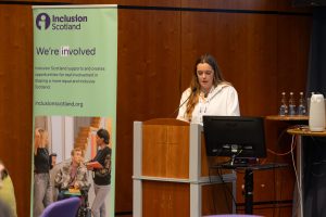 A speaker stood at the podium in a white shirt. There is an Inclusion Scotland roller banner to the left of the speaker.