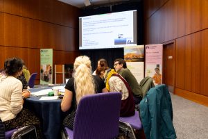 A group of attendees sitting at a round table at the Disability-inclusive climate emergency planning conference. There is a projector screen at the top of the image.