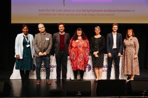 A group of seven speakers and candidates stand together on stage at Disabled People’s Question Time, smiling towards the audience.